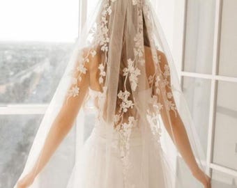 May include: A white bridal veil with delicate floral lace appliques. The veil is made of soft tulle and has a single-tier design.