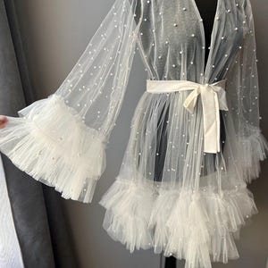 May include: A white sheer robe with pearl embellishments and a ruffled hem. The robe has a tie belt and long sleeves.
