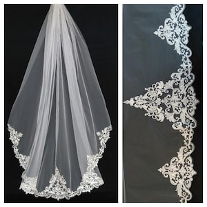 Op de afbeelding: Een witte bruids sluier met een delicate bloemenkant rand. De kant is zilverkleurig en heeft een delicaat, ingewikkeld ontwerp.