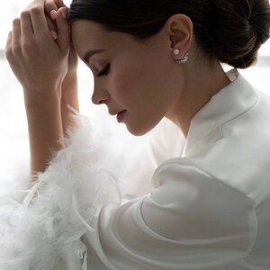 May include: A woman wearing a white satin robe with feather trim on the sleeves. She has her hair pulled back in a bun and is wearing pearl earrings.
