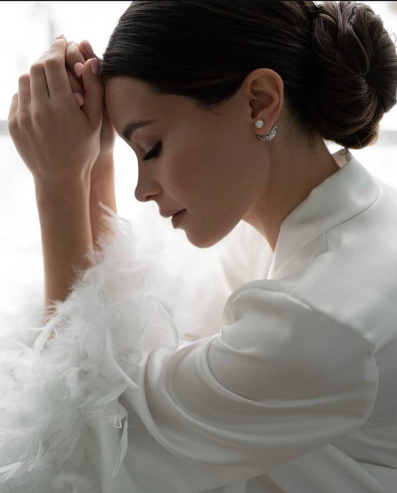 May include: A woman wearing a white satin robe with feather trim on the sleeves. She has her hair pulled back in a bun and is wearing pearl earrings.