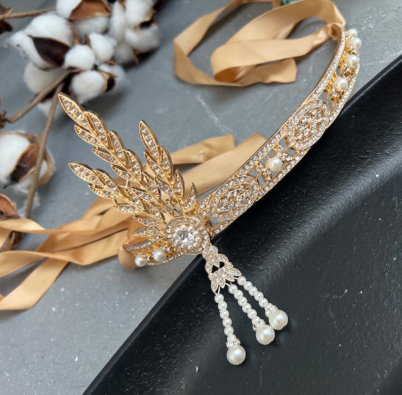 May include: A gold headband with a delicate, leaf-shaped design and a row of pearls. The headband has a dangling pearl drop with three strands of pearls.
