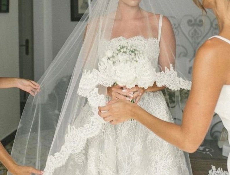 May include: A bride wearing a white lace wedding dress with a long veil. She is holding a bouquet of white flowers and is being helped by another person.