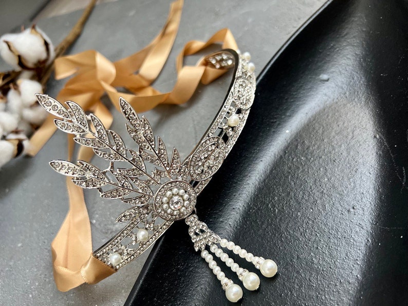 May include: A silver headband with a delicate floral design and pearl accents. The headband is adorned with rhinestones and has a dangling pearl chain.