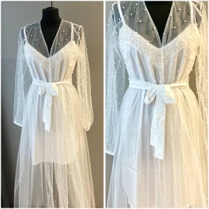 May include: White sheer maxi dress with pearl embellishments and a satin sash tie. The dress has long sleeves and a V-neckline.