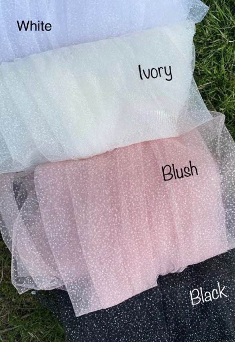 May include: Four swatches of sparkly tulle fabric in white, ivory, blush, and black. The fabric is layered with the white swatch on top and the black swatch on the bottom.