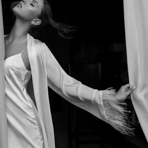 May include: A woman in a white satin robe with feather trim on the sleeves. She is standing in front of a white curtain and looking up.