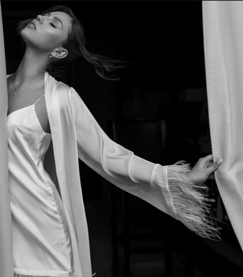 May include: A woman in a white satin robe with feather trim on the sleeves. She is standing in front of a white curtain and looking up.