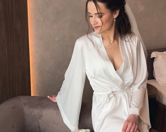 May include: A bride wearing a white satin robe with a white veil. She is sitting on a couch with a bouquet of white flowers and a small white purse.