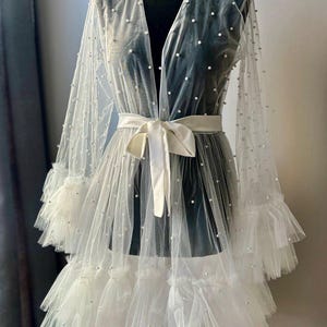 May include: A white sheer robe with a ruffled hem and pearl embellishments. The robe has a satin sash and long sleeves.