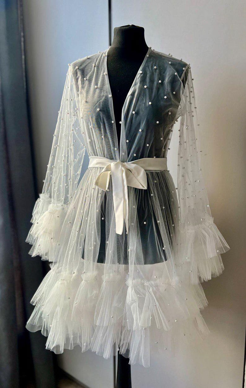 May include: A white sheer robe with a ruffled hem and pearl embellishments. The robe has a satin sash and long sleeves.