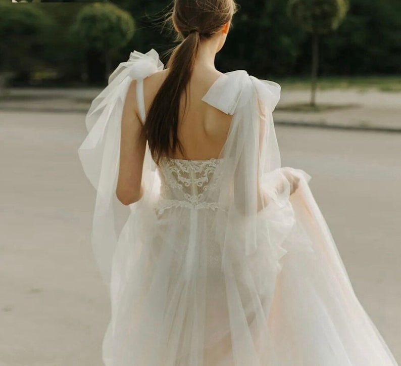 May include: A white tulle wedding dress with a lace bodice and large bow details on the straps. The dress has a flowing skirt and a long train.