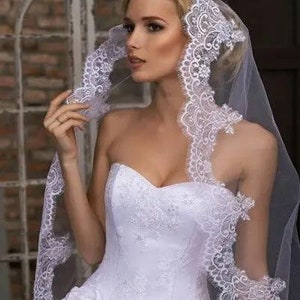 May include: A white bridal veil with a delicate lace trim. The veil is made of soft tulle and has a single layer. The lace trim is intricate and features a floral design.