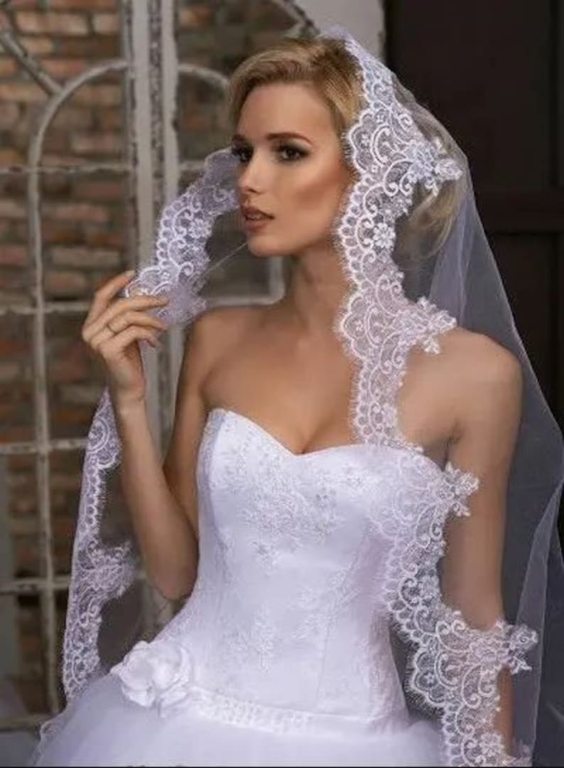 May include: A white bridal veil with a delicate lace trim. The veil is made of soft tulle and has a single layer. The lace trim is intricate and features a floral design.