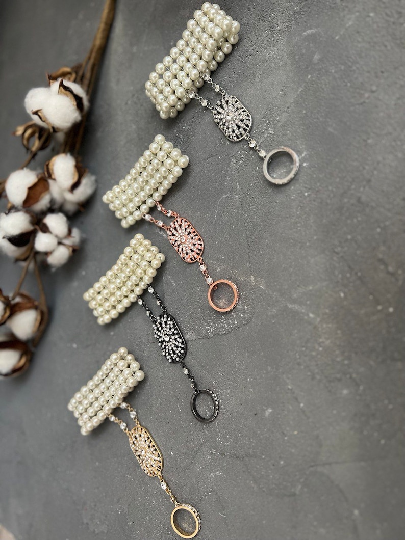 May include: Four pearl bracelets with a rhinestone ring attached to each. The bracelets are white and the rings are silver, rose gold, black, and gold.