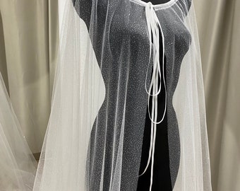 May include: White tulle cape with a sparkly finish and a white satin tie closure. The cape is sheer and has a delicate, flowing design.