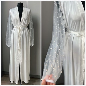 May include: White satin robe with a long, flowing silhouette and a tie belt. The robe has sheer, pearl-embellished sleeves.