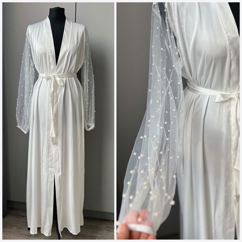 May include: White satin robe with a long, flowing silhouette and a tie belt. The robe has sheer, pearl-embellished sleeves.