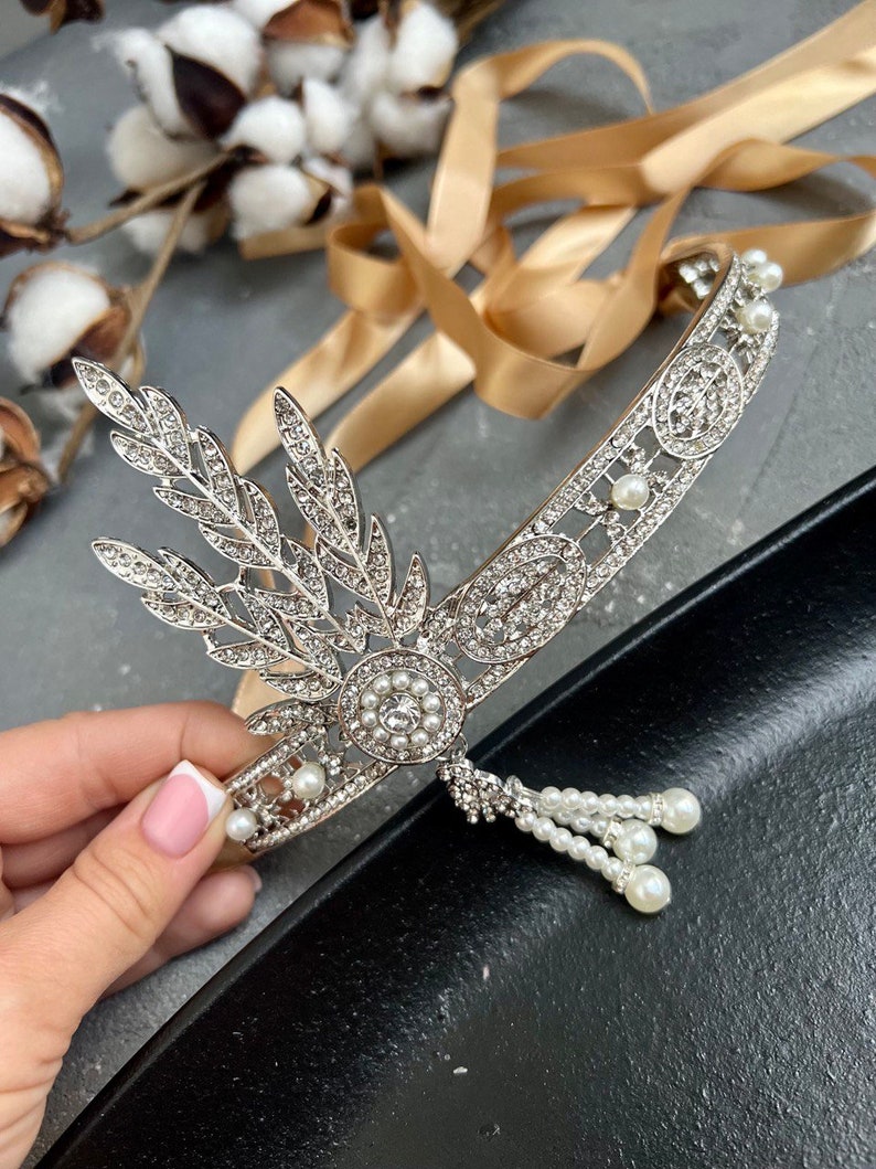 May include: A silver headband with a pearl and rhinestone design. The headband has a leaf-like design on one side and a pearl drop on the other.