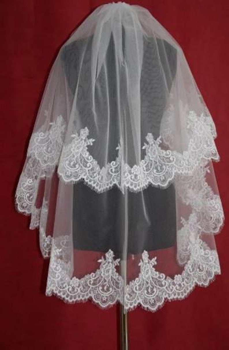 May include: A white bridal veil with three layers of lace trim. The veil is made of soft tulle and features delicate floral patterns.