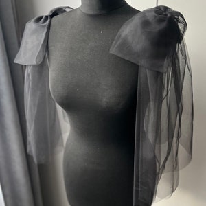 May include: Black tulle shoulder pads with large bows on each shoulder. The shoulder pads are on a black dress form.