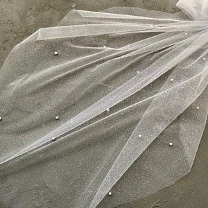 May include: A white tulle veil with small pearl embellishments. The veil is folded in half and has a delicate, sparkly texture.