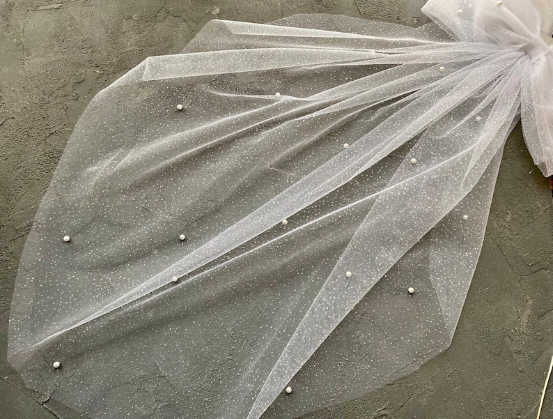 May include: A white tulle veil with small pearl embellishments. The veil is folded in half and has a delicate, sparkly texture.