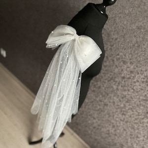 May include: A white tulle bridal veil with a large white bow adorned with pearl-like embellishments. The veil is draped over a black dress form.