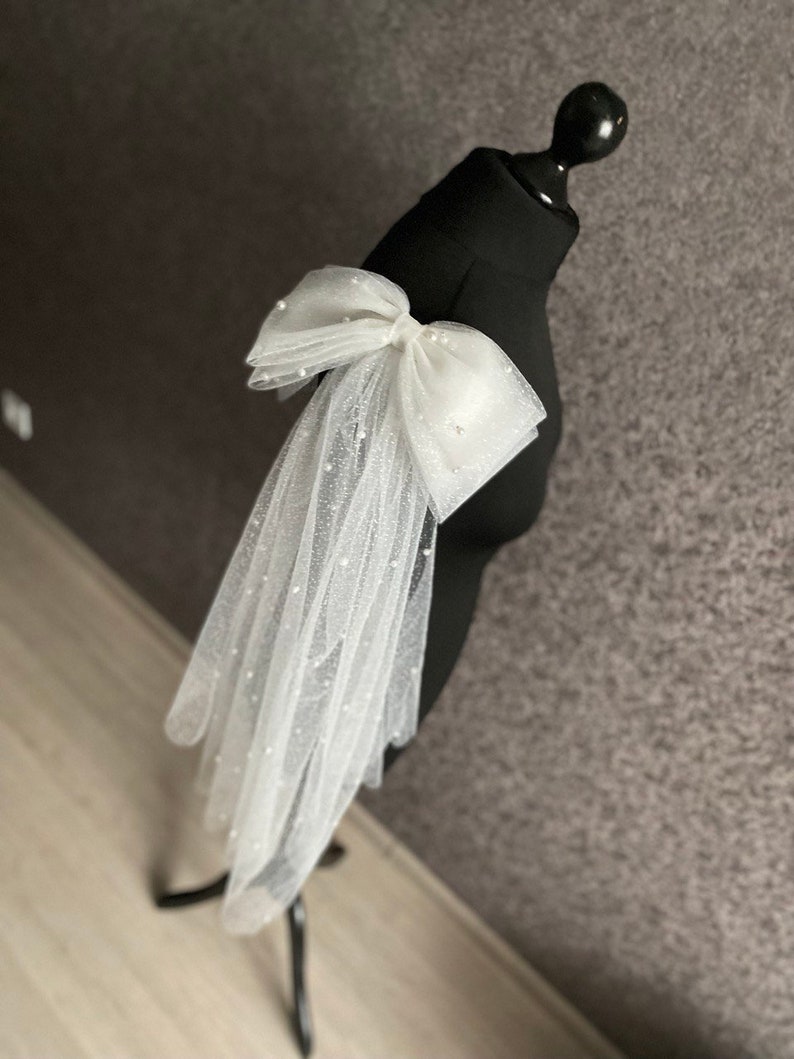 May include: A white tulle bridal veil with a large white bow adorned with pearl-like embellishments. The veil is draped over a black dress form.