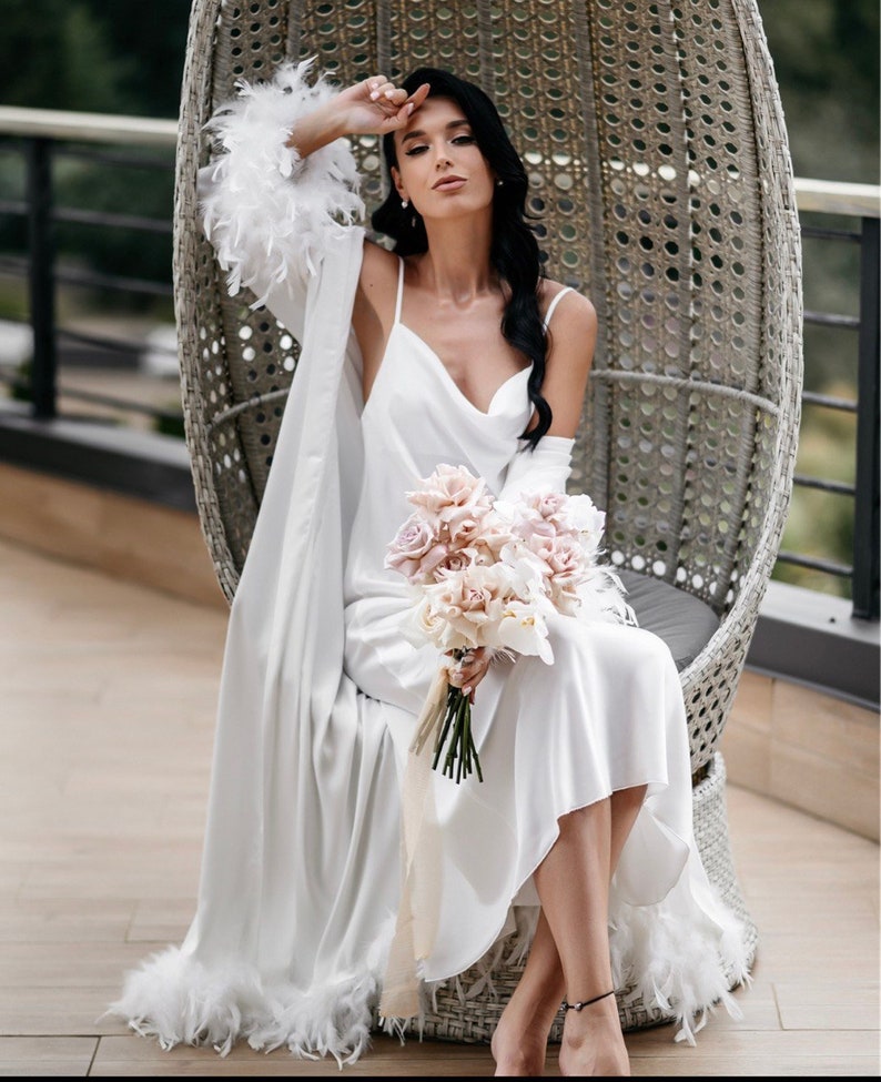 May include: A white satin robe with feather trim, worn over a white satin slip dress. The person is holding a bouquet of pink and white flowers.