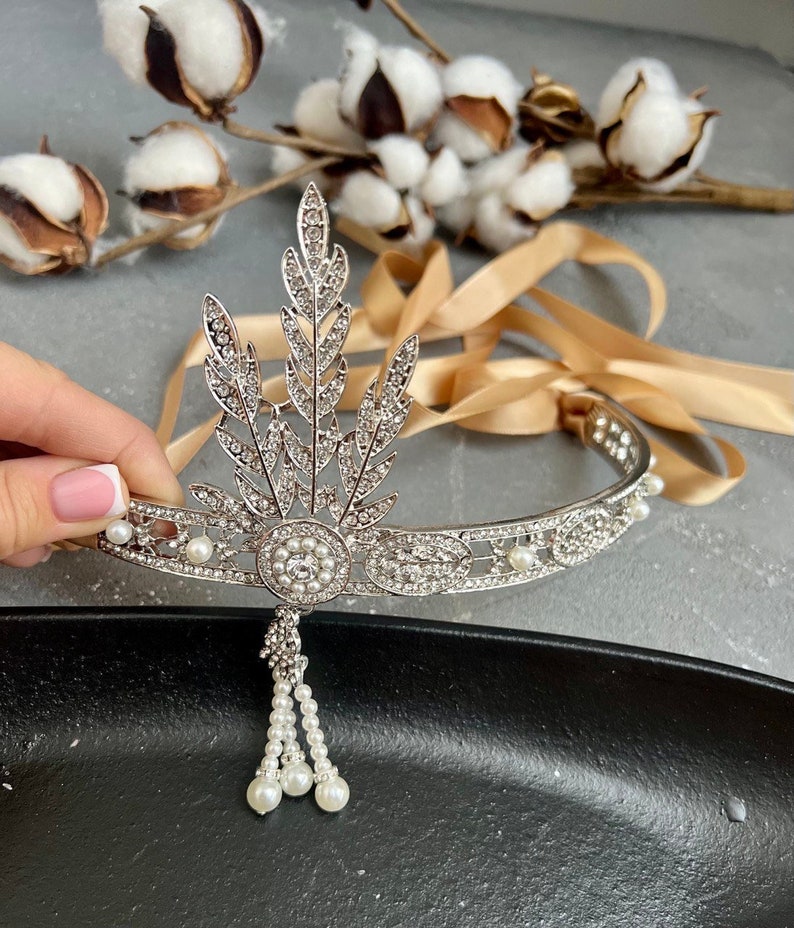May include: A silver headband with a pearl and crystal design. The headband has a large, ornate, leaf-shaped design in the front with a dangling pearl tassel.