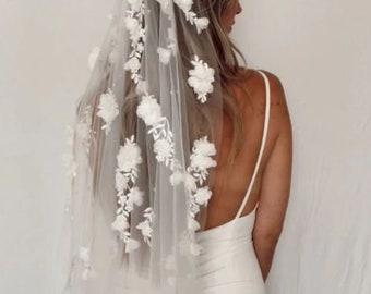 May include: A white bridal veil with delicate floral embellishments. The veil is made of soft tulle and features a delicate floral pattern throughout. The veil is worn by a person with long brown hair.