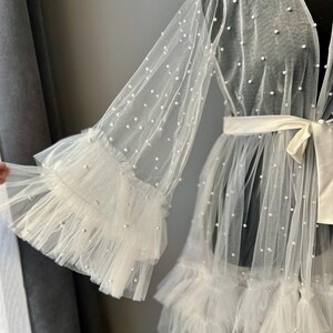 May include: A white sheer robe with pearl embellishments and ruffled trim. The robe has long sleeves and a tie at the waist.