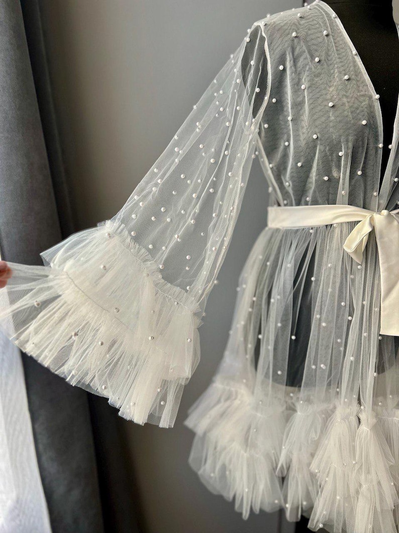 May include: A white sheer robe with pearl embellishments and ruffled trim. The robe has long sleeves and a tie at the waist.