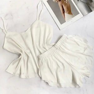 Puede incluir: Conjunto de pijama de satén blanco con top de tirantes finos y pantalones cortos. El top tiene cuello en V y los pantalones cortos tienen un dobladillo con volantes.