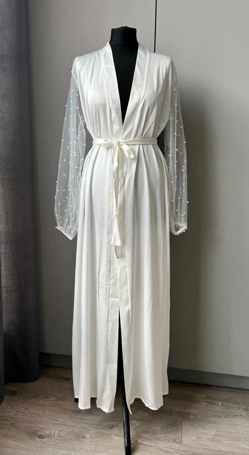 May include: A white satin robe with a long, flowing skirt and a sash tie at the waist. The sleeves are made of sheer mesh with pearl-like embellishments.