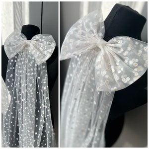May include: A white tulle bridal veil with a large white bow decorated with small white daisies. The veil is made of soft, delicate fabric and has a romantic, whimsical feel.