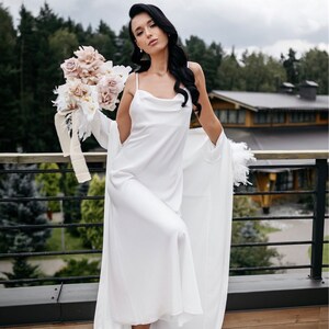 May include: A white satin slip dress with a cowl neck and feather trim. The dress is worn with a white feather robe. The model is holding a bouquet of blush pink and white flowers.