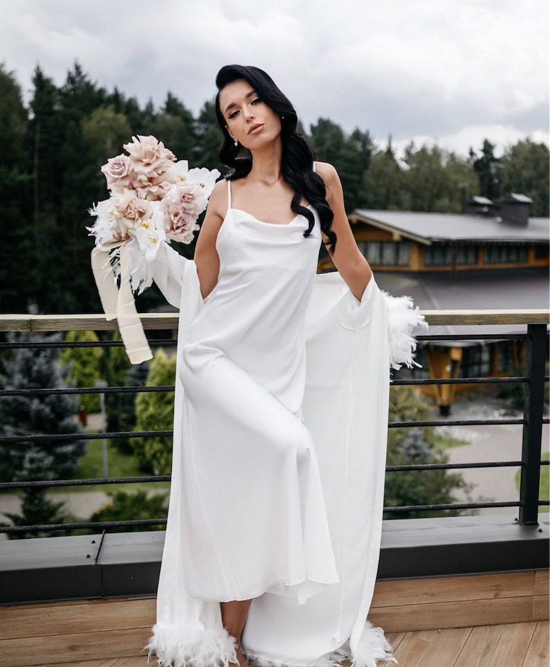 May include: A white satin slip dress with a cowl neck and feather trim. The dress is worn with a white feather robe. The model is holding a bouquet of blush pink and white flowers.