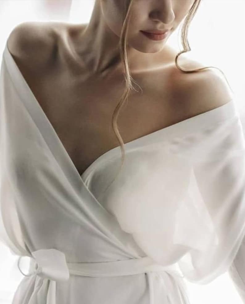 May include: A white satin robe with a wrap style and a tie at the waist. The robe has long sleeves and a deep V-neckline.