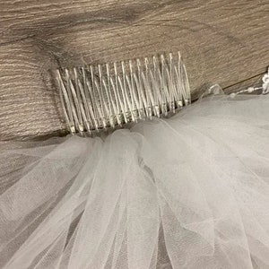 May include: A white tulle bridal veil with a comb attached to the top. The veil has a lace trim around the edge.