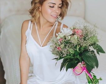 Pode incluir: Uma mulher com um vestido branco de renda segura um buquê de flores brancas, rosas e verdes. O buquê está amarrado com uma fita rosa.