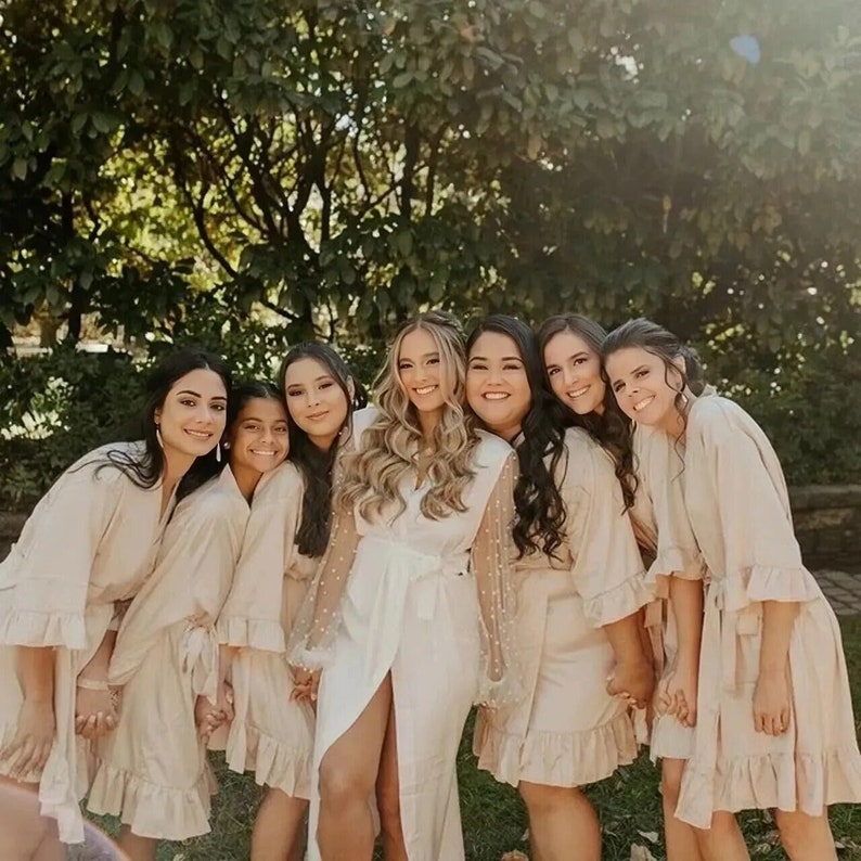 May include: A group of seven women wearing white and beige robes. The woman in the center is wearing a white robe with pearl-embellished sleeves. The other women are wearing beige robes with ruffled sleeves. The women are smiling and looking at the camera.