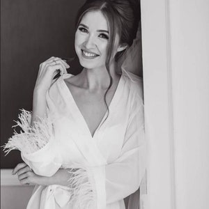 May include: A woman in a white robe with feather trim on the sleeves. She is smiling and looking at the camera. The robe is sheer and has a V-neckline.