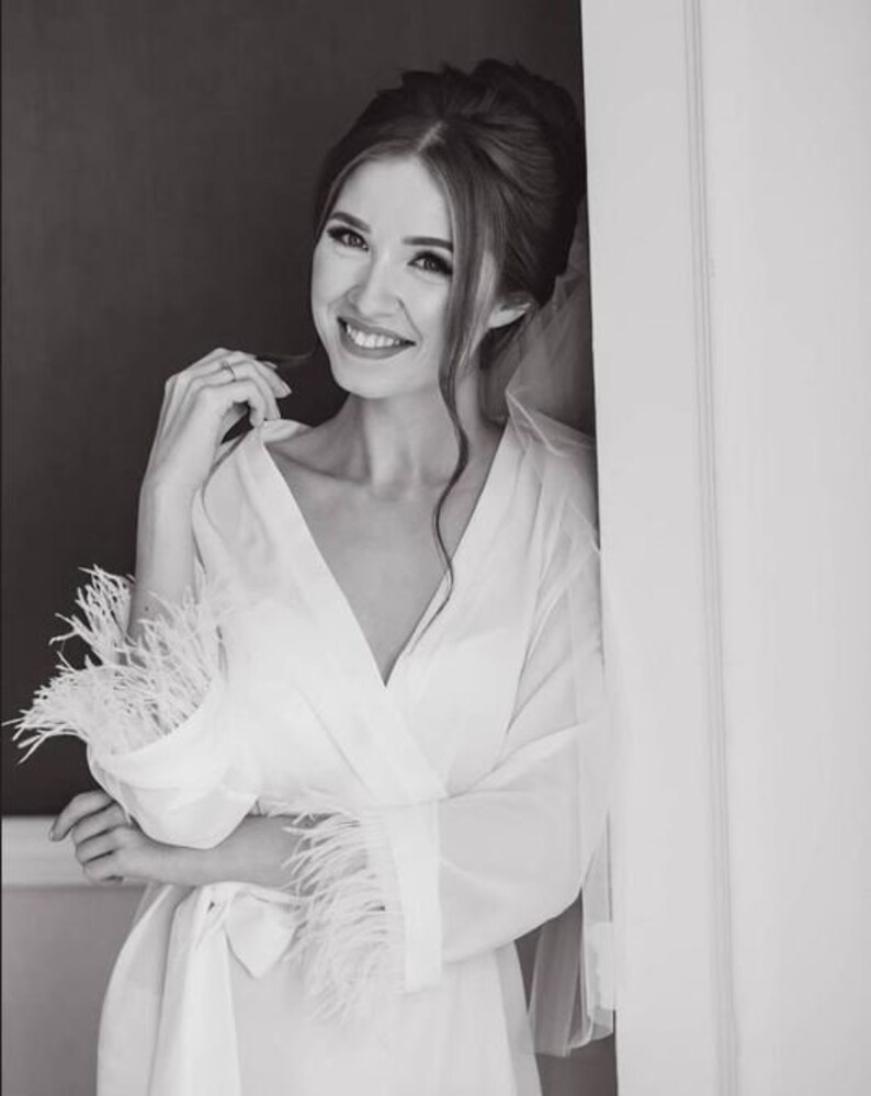 May include: A woman in a white robe with feather trim on the sleeves. She is smiling and looking at the camera. The robe is sheer and has a V-neckline.