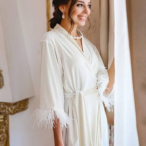 May include: A white velvet robe with feather trim on the sleeves and a sash tie. The robe is worn by a person with a pearl necklace and earrings. The person is holding a bouquet of pink flowers.