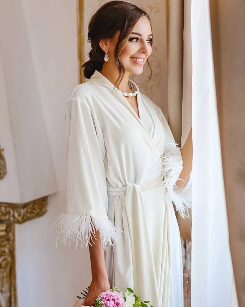 May include: A white velvet robe with feather trim on the sleeves and a sash tie. The robe is worn by a person with a pearl necklace and earrings. The person is holding a bouquet of pink flowers.