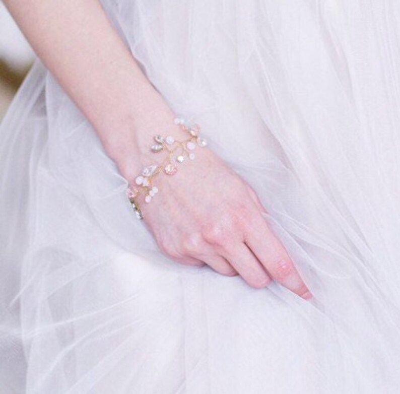May include: A delicate gold bracelet with pink and white crystals arranged in a floral design. The bracelet is made of wire and has a delicate, feminine look.