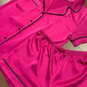 May include: A pink satin pajama set with black trim. The top has short sleeves and a button-up front. The shorts have a drawstring waist.