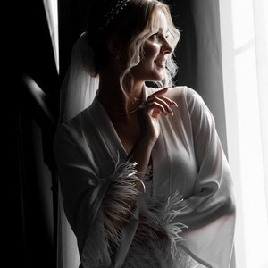 May include: A bride in a white satin robe with feather trim, standing by a window. She is wearing a veil and a tiara.